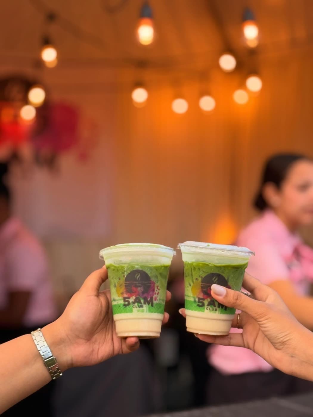 Two hands hold up matching iced drinks in cups printed with the 5AM Club logo against a string-light backdrop.
