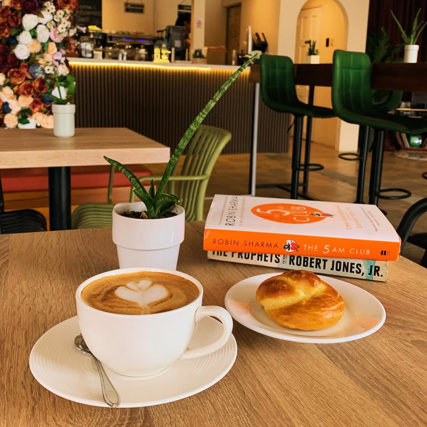 A latte with heart latte art, a croissant, and a copy of 'The 5 AM Club' book arranged on a wooden table in the cafe seating area.