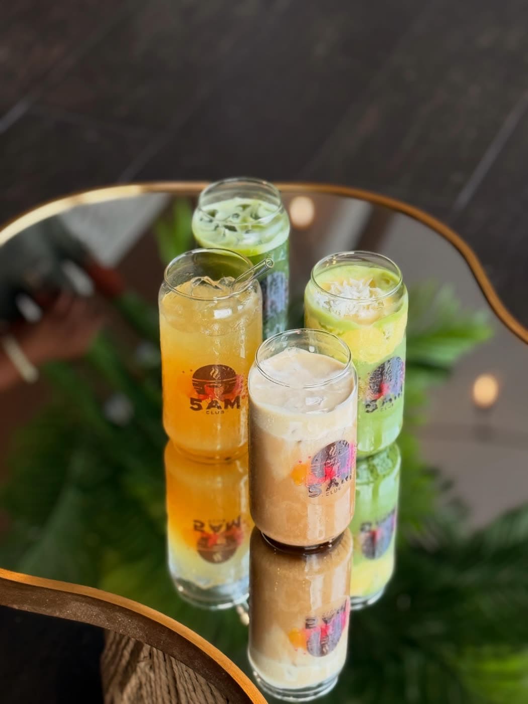 Five different colored iced drinks are arranged closely on a mirrored table reflecting their glasses.