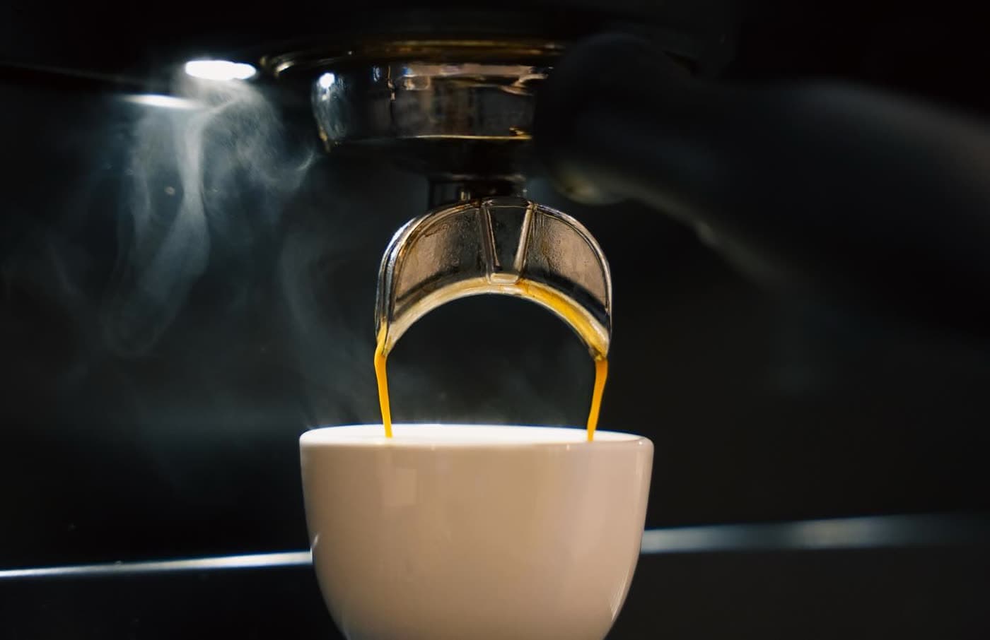 Espresso streaming from a portafilter into a white cup with visible steam against a dark background.