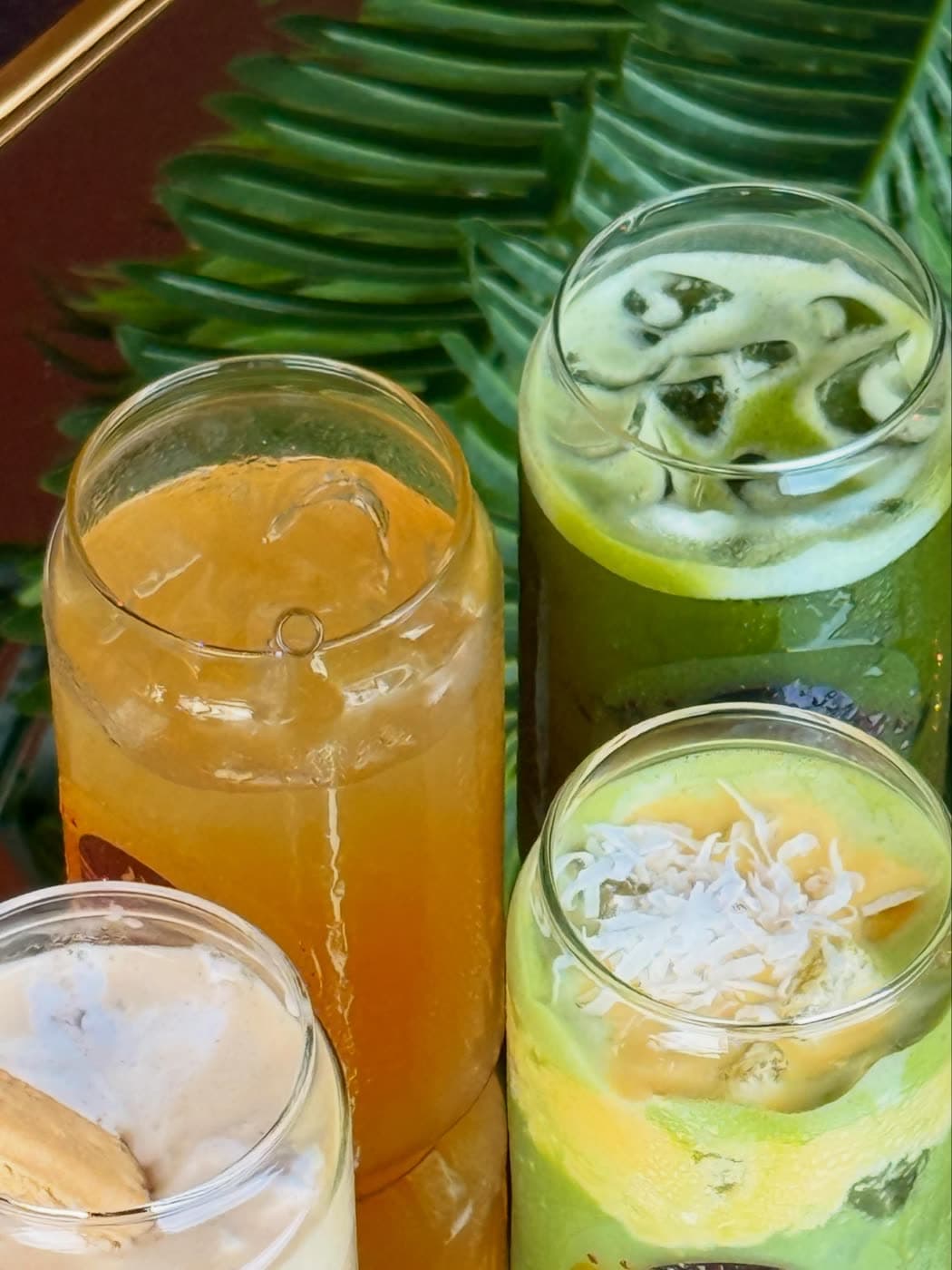 Four iced drinks in different colors are grouped closely together beside a large palm leaf.