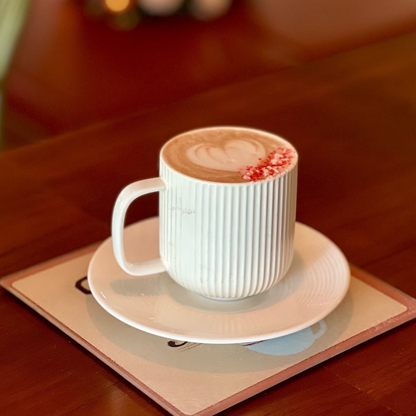 A cappuccino with heart-shaped latte art and a dusting of red sprinkles, served in a white ribbed mug on a saucer.
