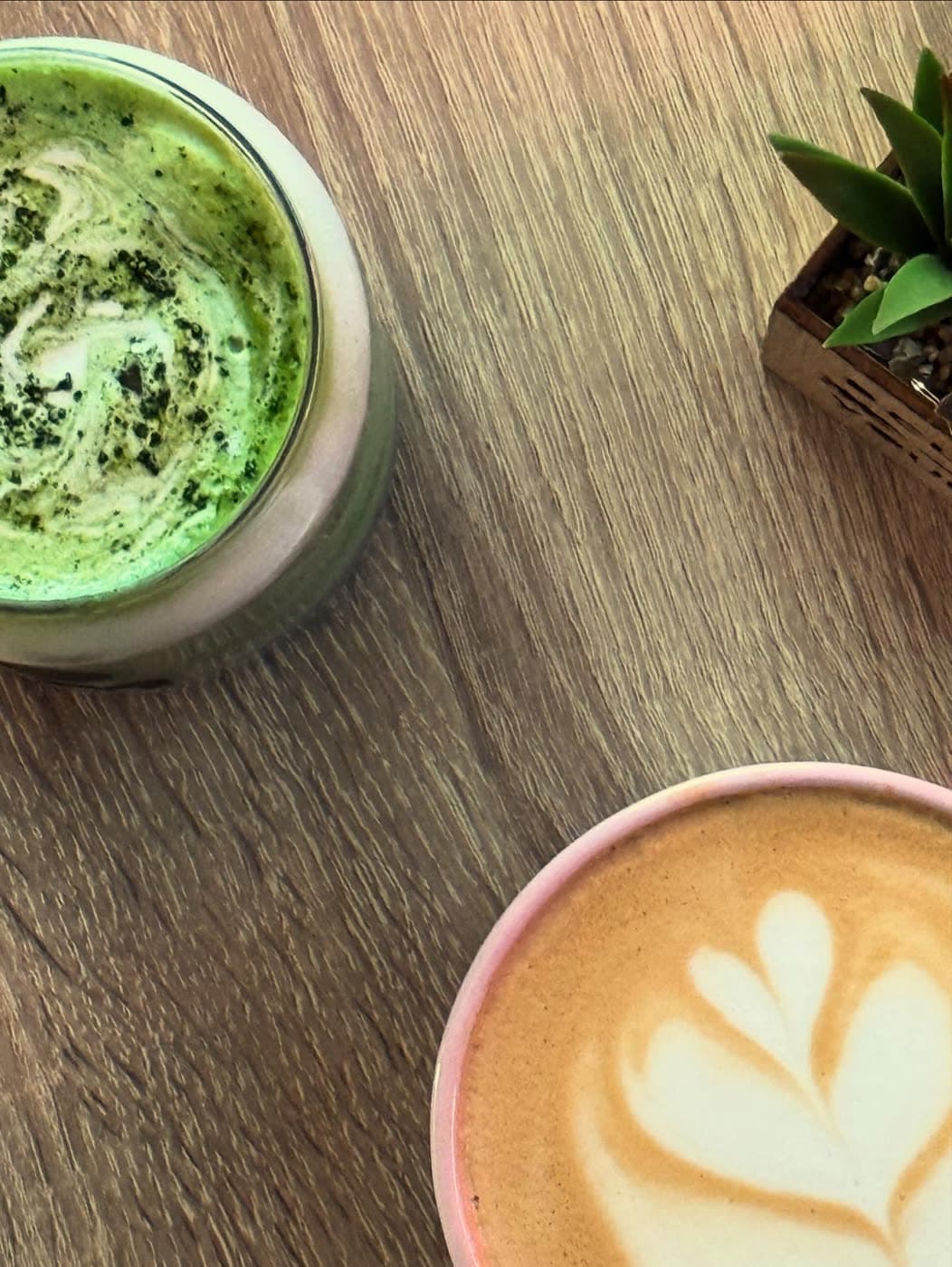 An overhead view of an iced matcha and a latte with heart art on a wood table beside a small plant.