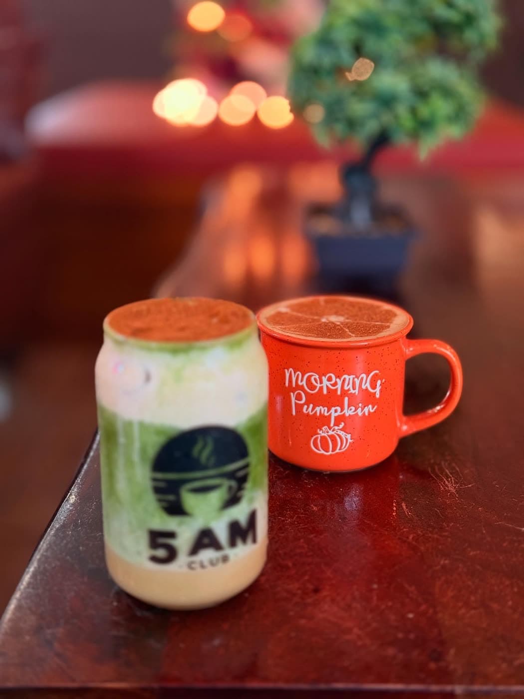 An iced matcha drink and an orange 'Morning Pumpkin' mug sit together on a table by candlelight.