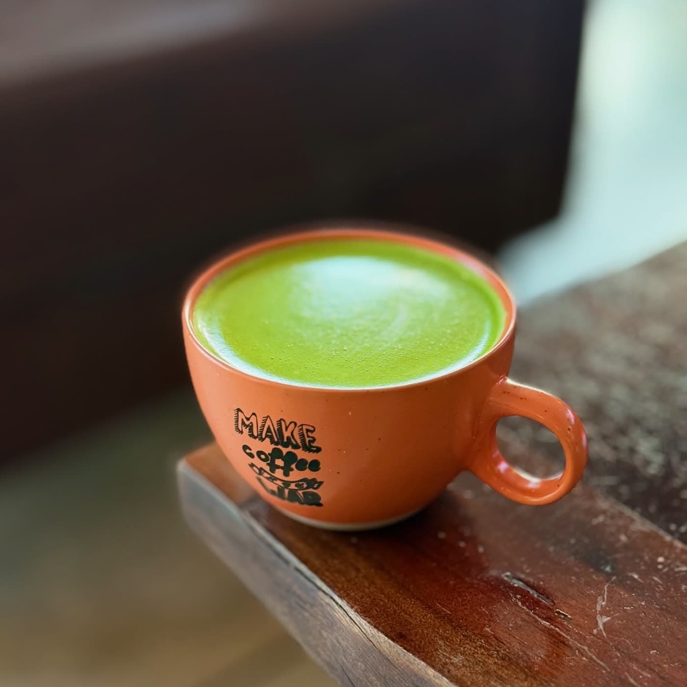 A matcha latte with a light foam swirl served in an orange cup on a wooden ledge.