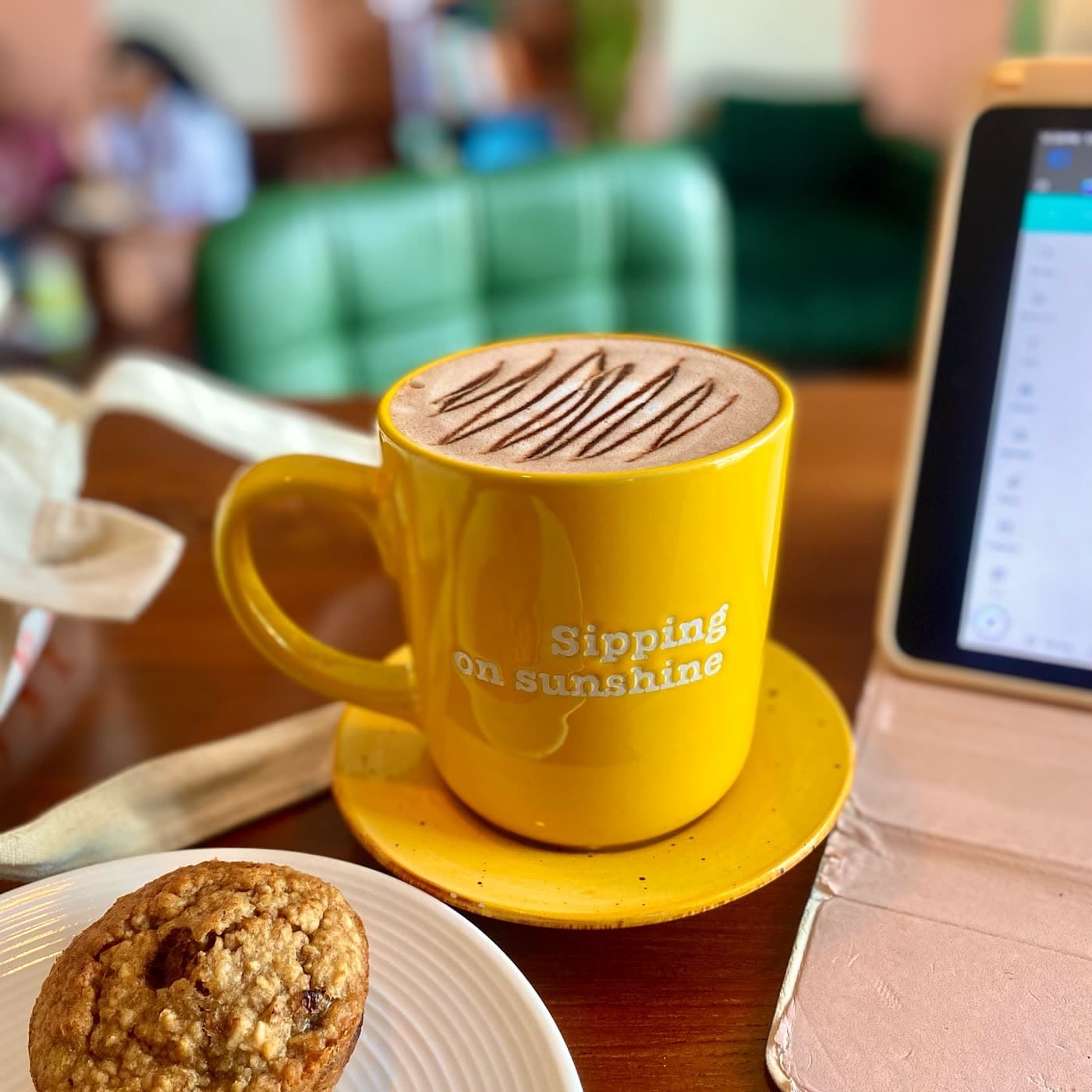 A yellow ceramic mug reading 'Sipping on sunshine' filled with latte art coffee, beside an oatmeal cookie on a wooden table.