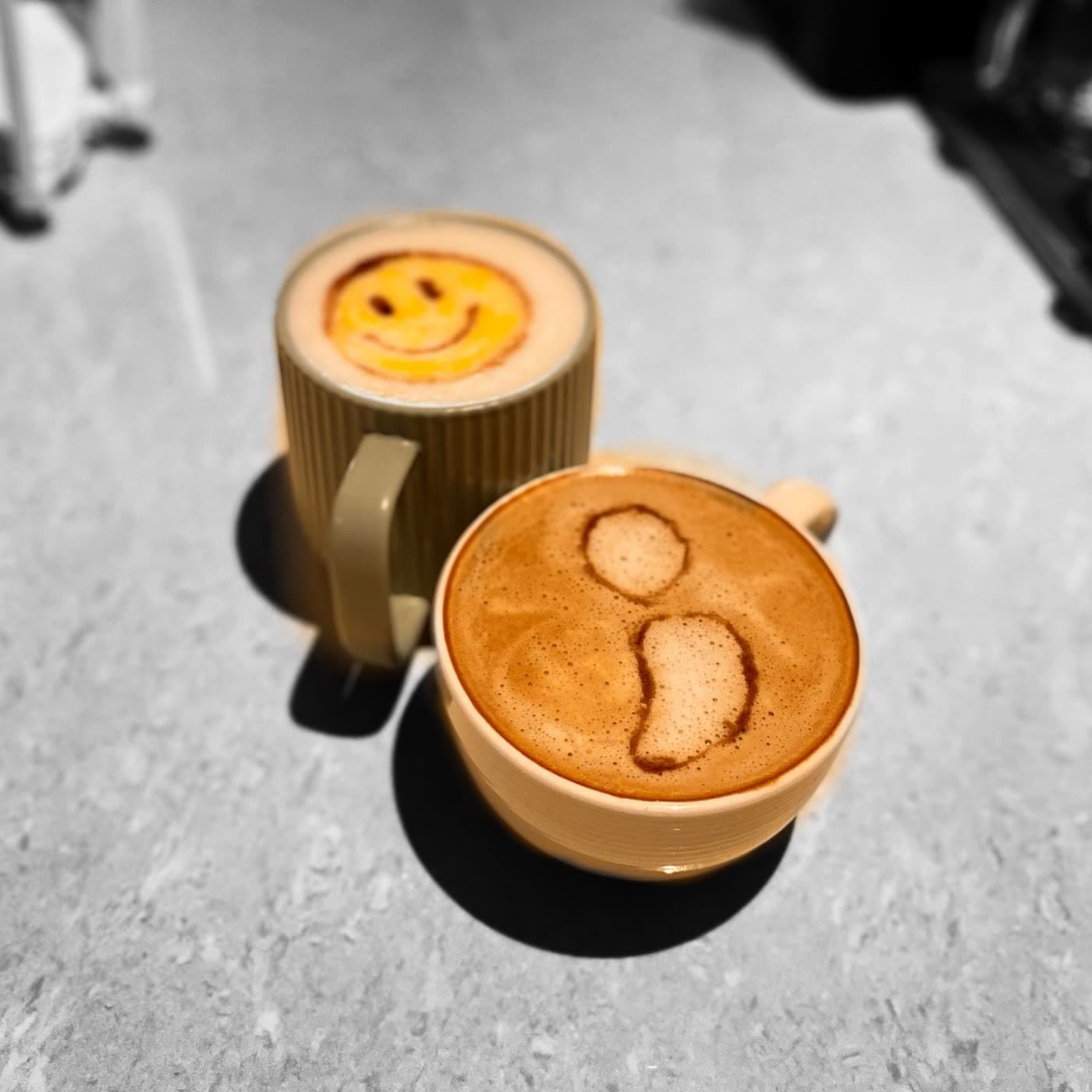 Two lattes side by side on a table, one with a smiley face and one with a frowning face drawn in the foam.