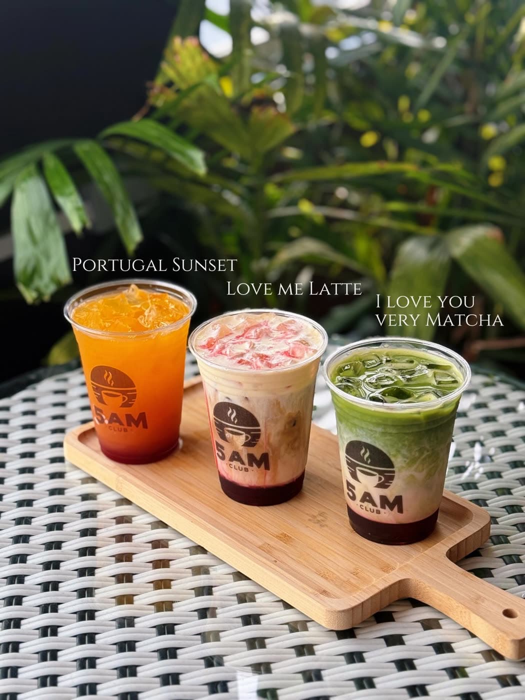 Three labeled iced drinks are arranged on a wooden paddle board against greenery.
