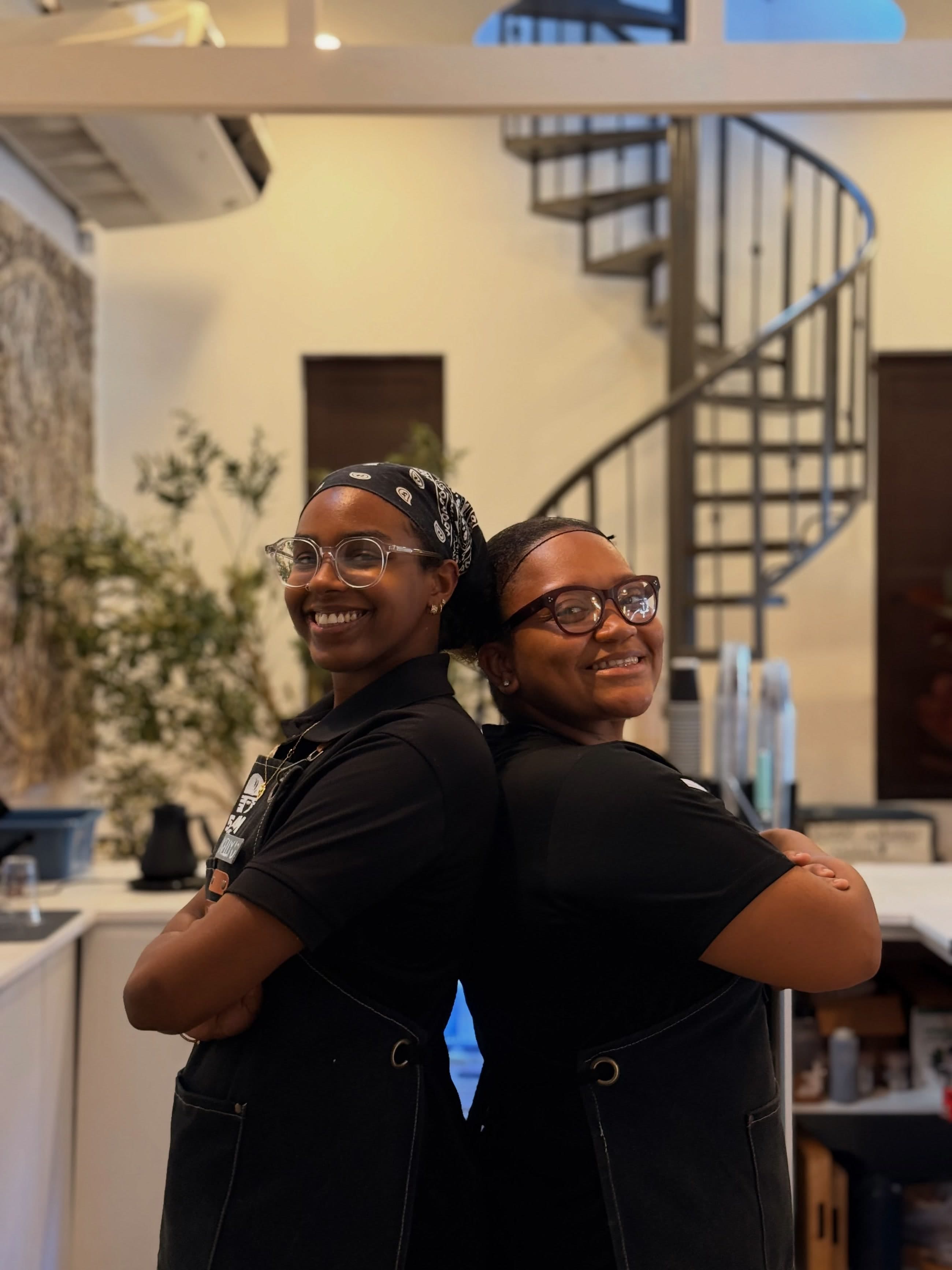 Two baristas stand back to back and smiling inside the loft, spiral staircase and plants behind them