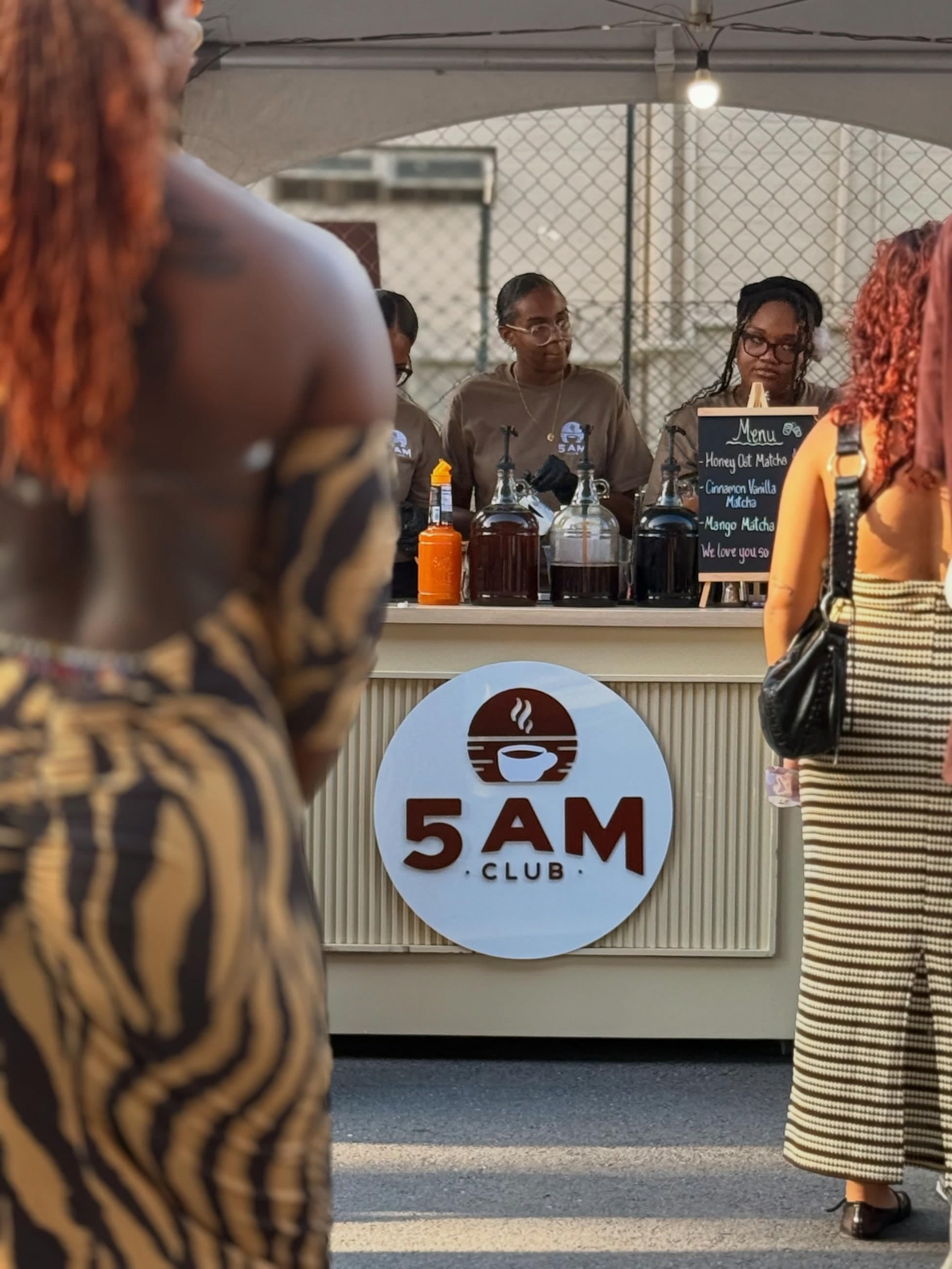 The 5AM Club cart seen from the front, logo panel lit, three staff pouring behind it while customers wait