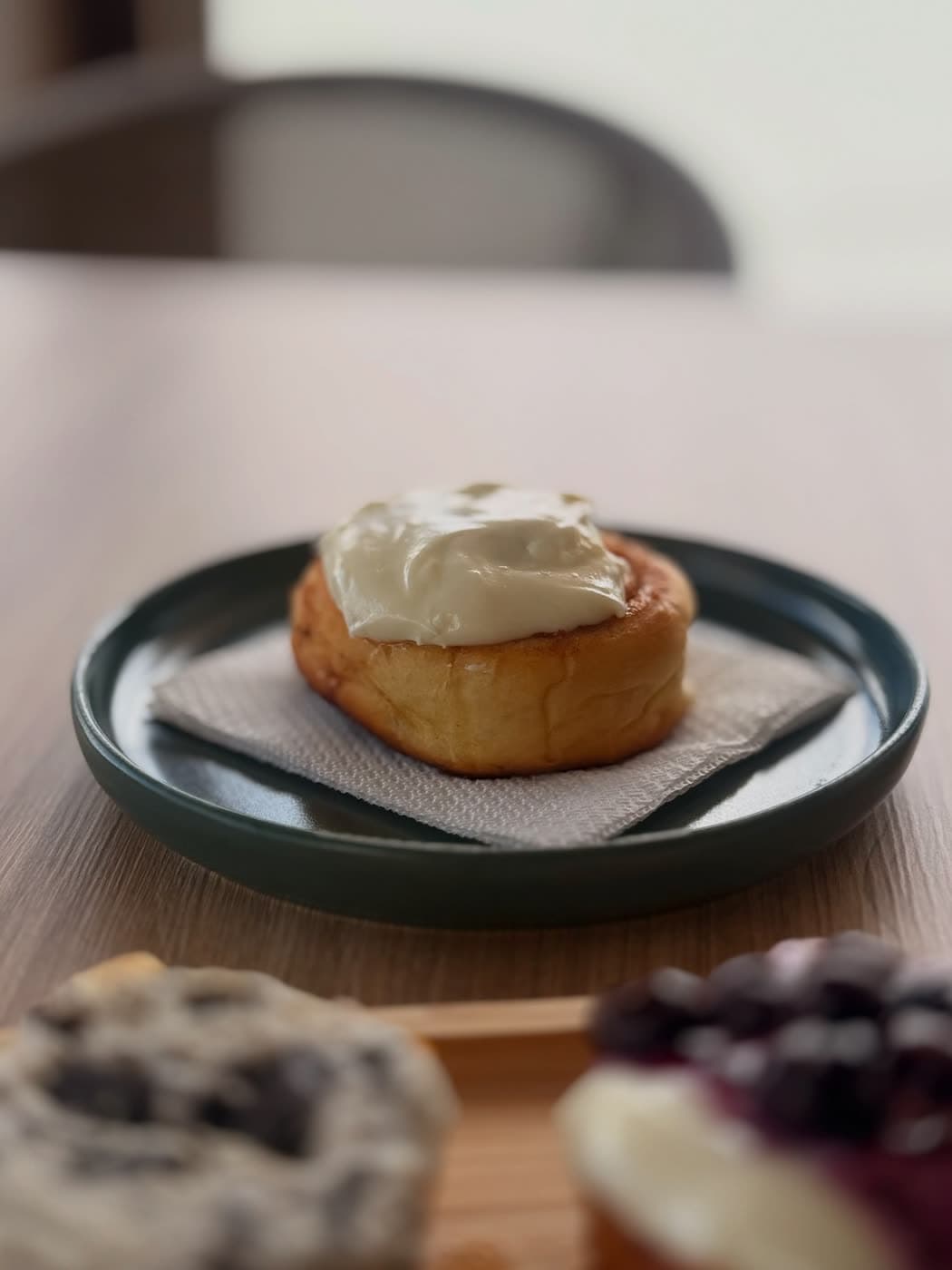 A cinnamon roll with cream cheese frosting on a dark green plate, pastries blurred in front