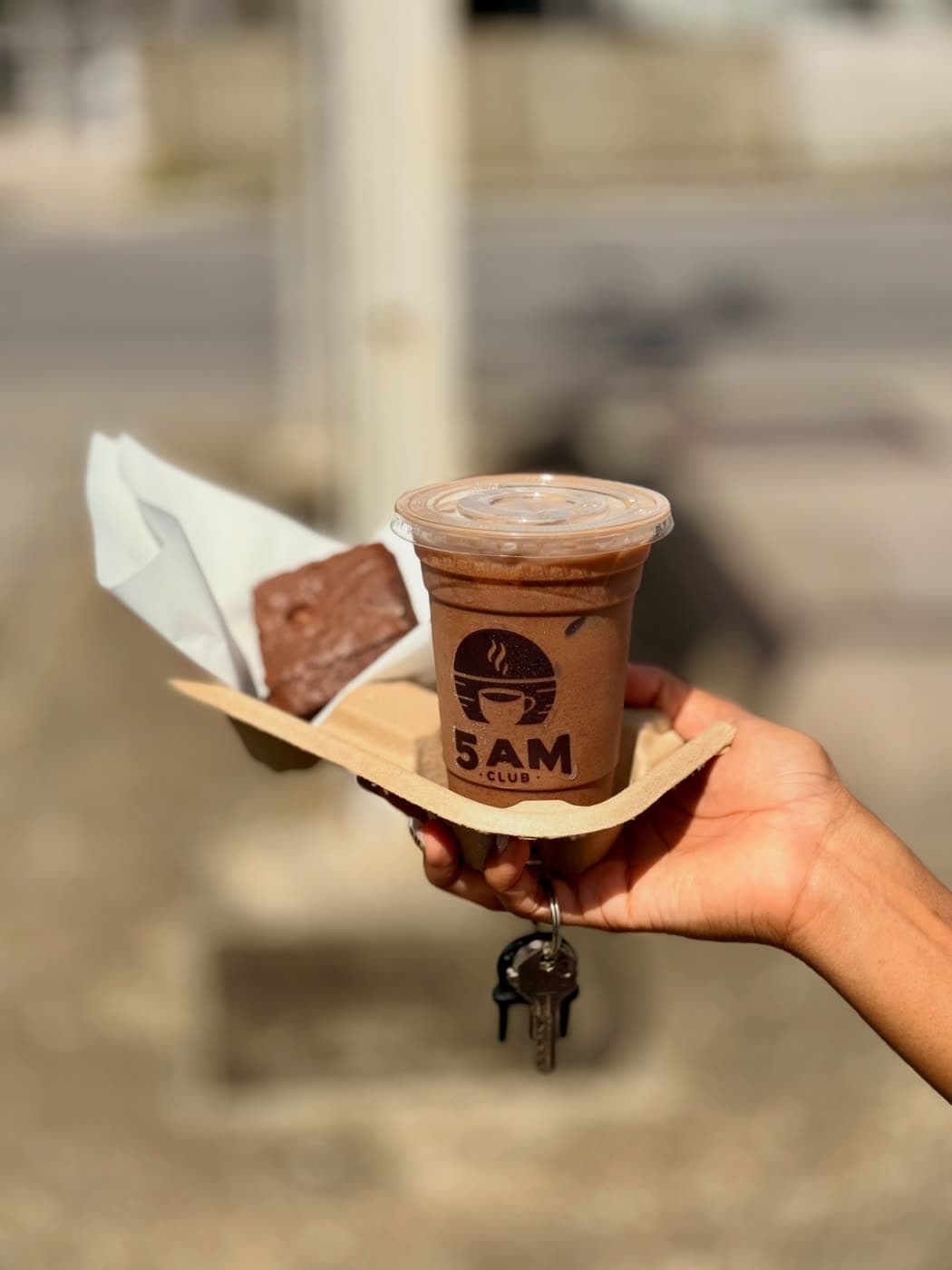 A hand holds a branded 5AM iced coffee and a brownie in a kraft carrier on a sunlit street