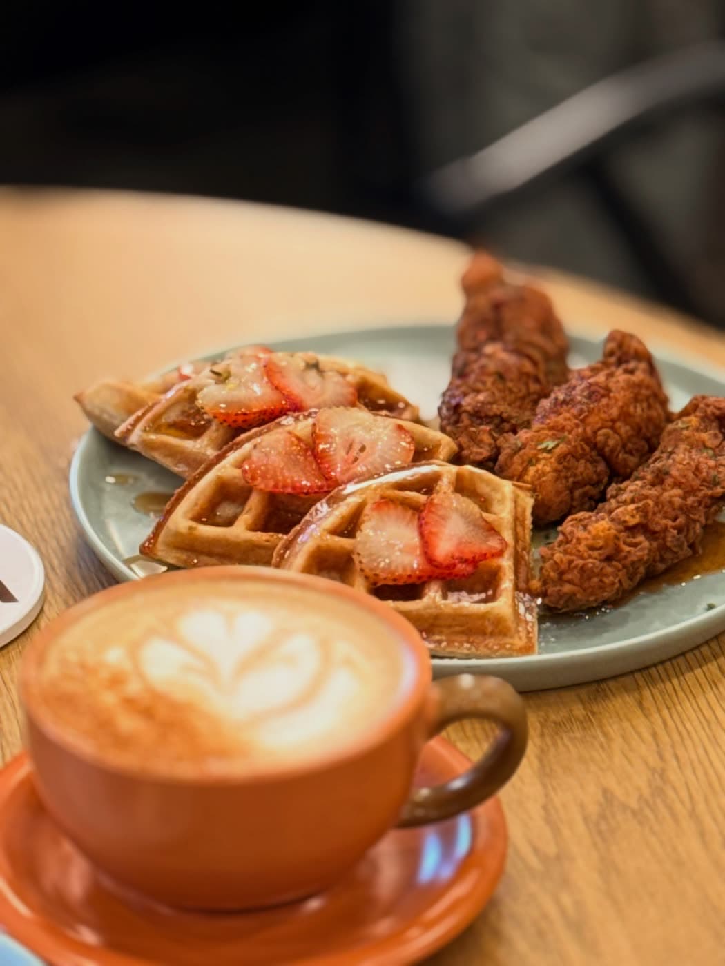 A rosetta latte in a terracotta cup with waffles and chicken softly out of focus behind