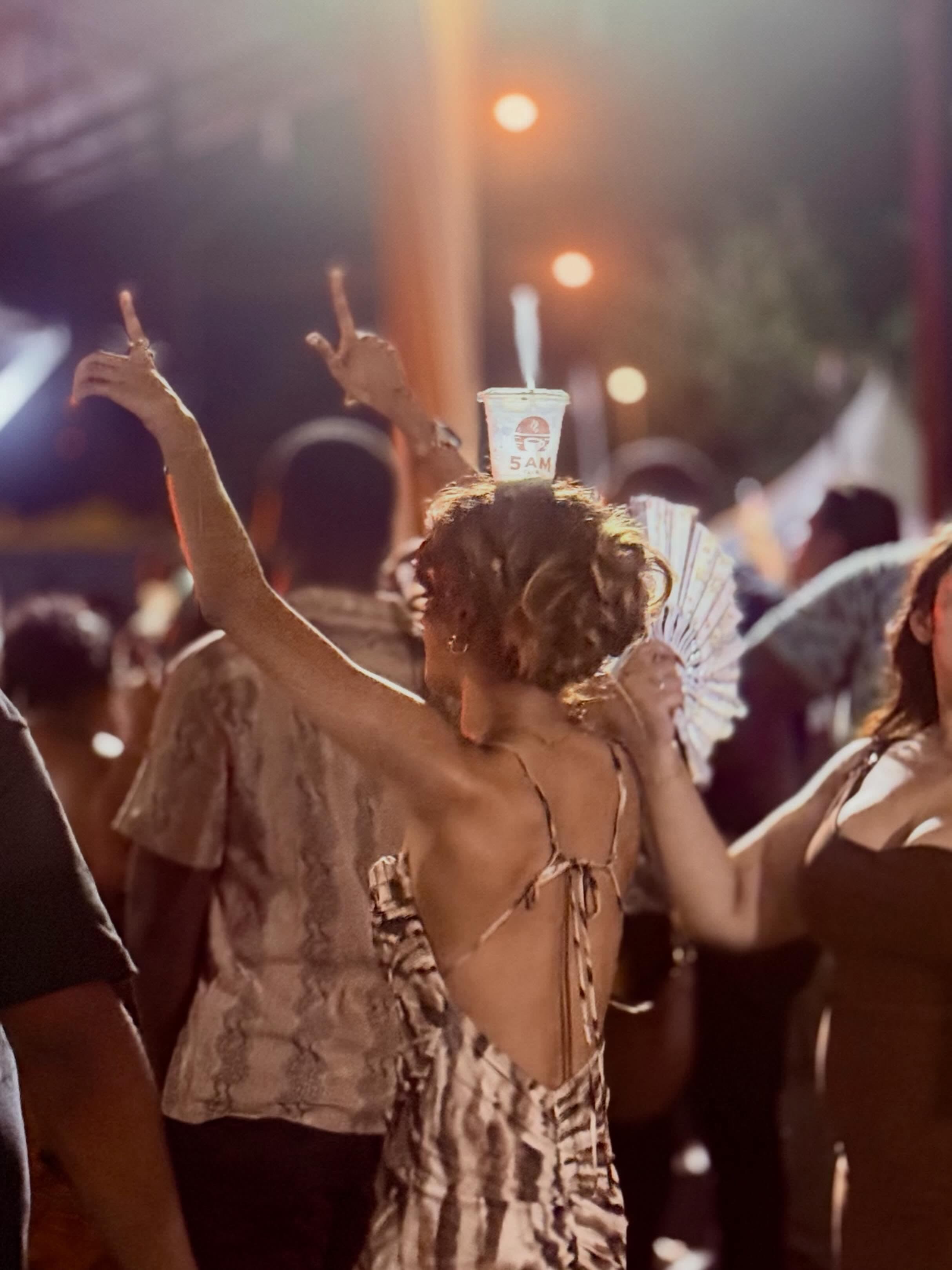 A dancer at a night market balances a 5AM Club cup on her head, hands raised, under warm string lights