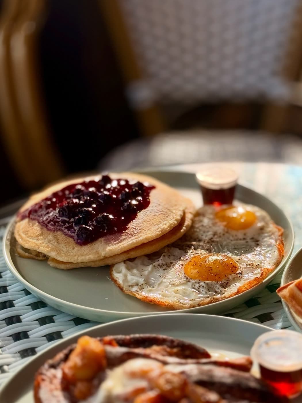 Pancakes with berry compote and two fried eggs beside a syrup cup in window light