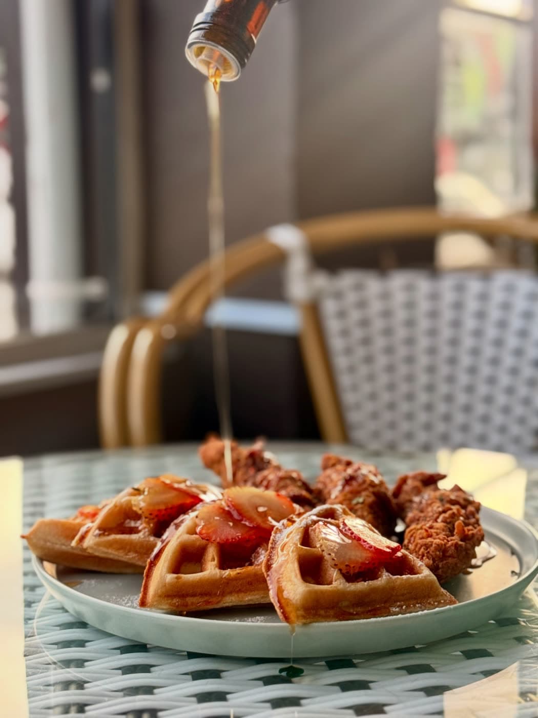 Syrup pours mid-air onto strawberry waffles and chicken tenders, café window and rattan chairs behind